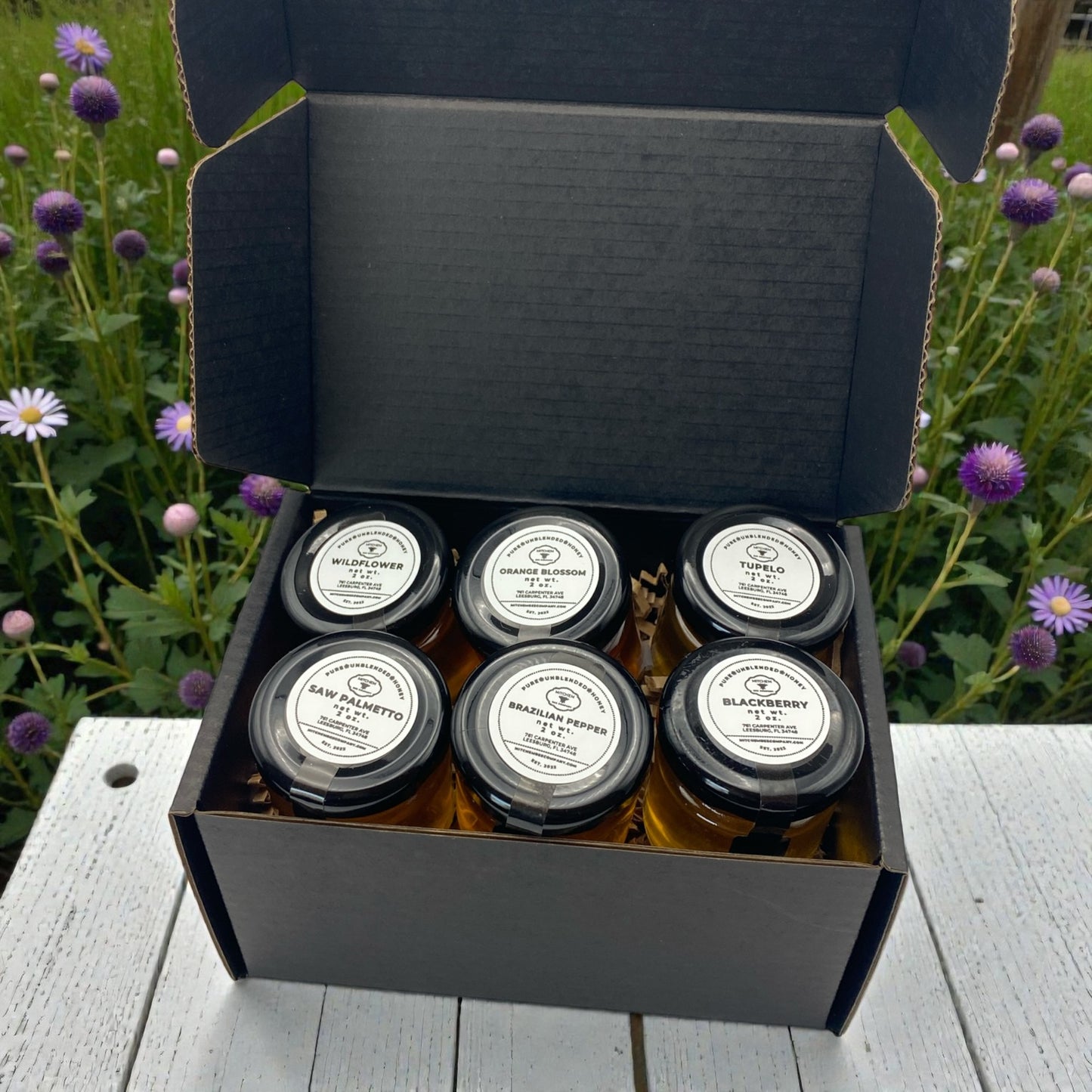 Box of six glass honey jars with labels on a wooden surface with flowers in the background