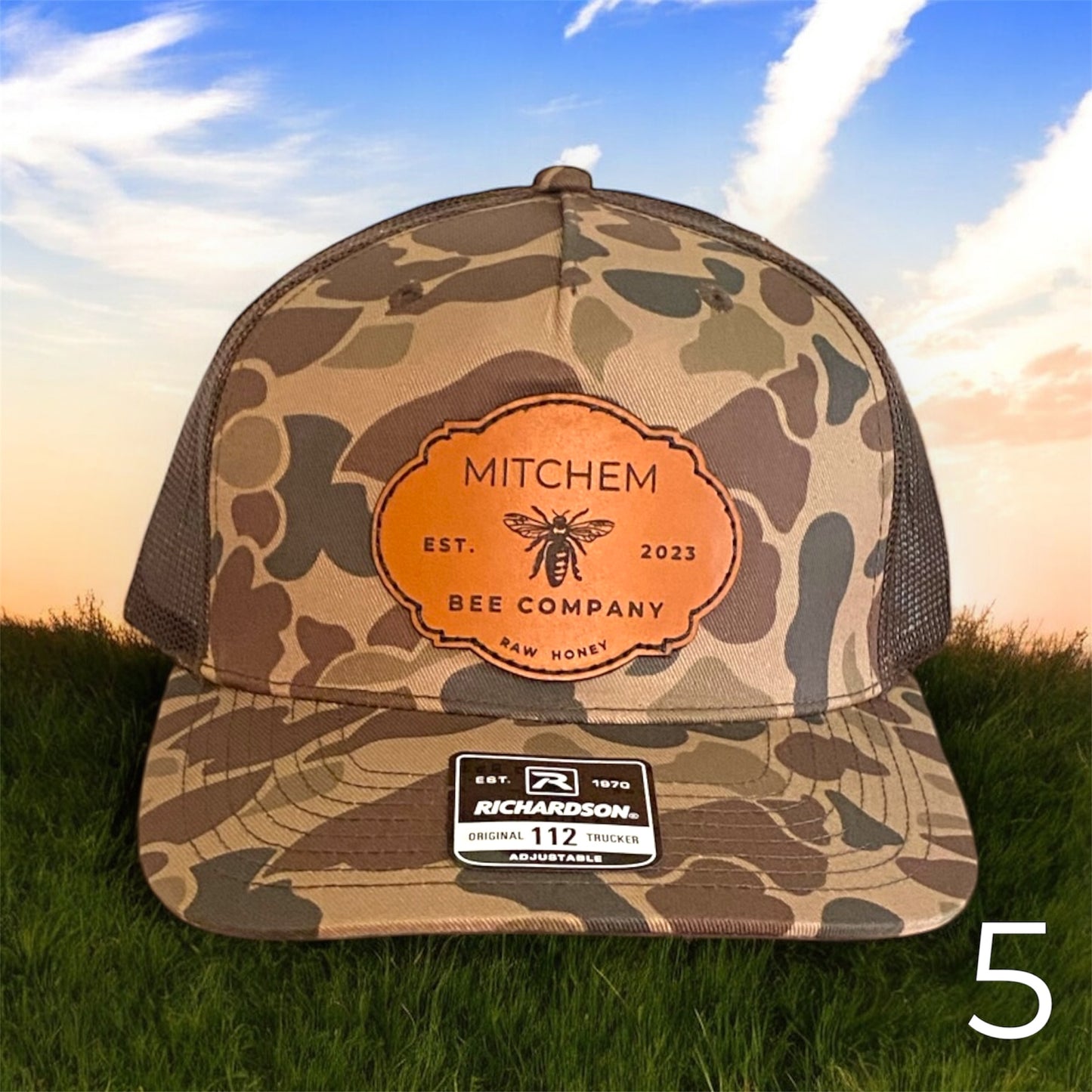 Brown Camouflage cap with a Mitchem Bee Company brown leather patch on grass with a sunset background