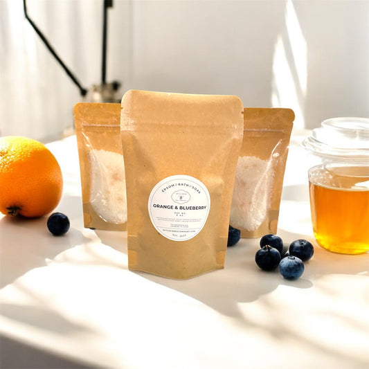 Three brown paper pouches of Epsom bath soaks labeled 'Orange & Blueberry' on a white surface with an orange, blueberries, and a glass of honey.