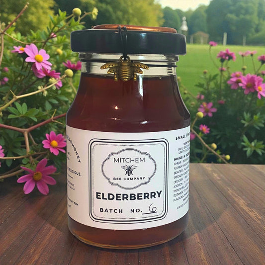 Jar of Mitchem Bee Company Elderberry honey with a bee decoration on a wooden table with flowers in the background.