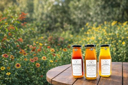 Three glass honey bottles labeled Wildflower Orange Blossom and Tupelo on a wooden table in daylight with wildflowers in the natural greenery background