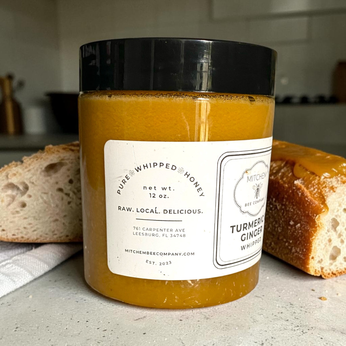 A jar of Kitchen Bee Company Pure Whipped Honey on a kitchen counter, flanked by slices of rustic bread. The jar has a black lid and a white label indicating the flavor is Turmeric Ginger Whipped.