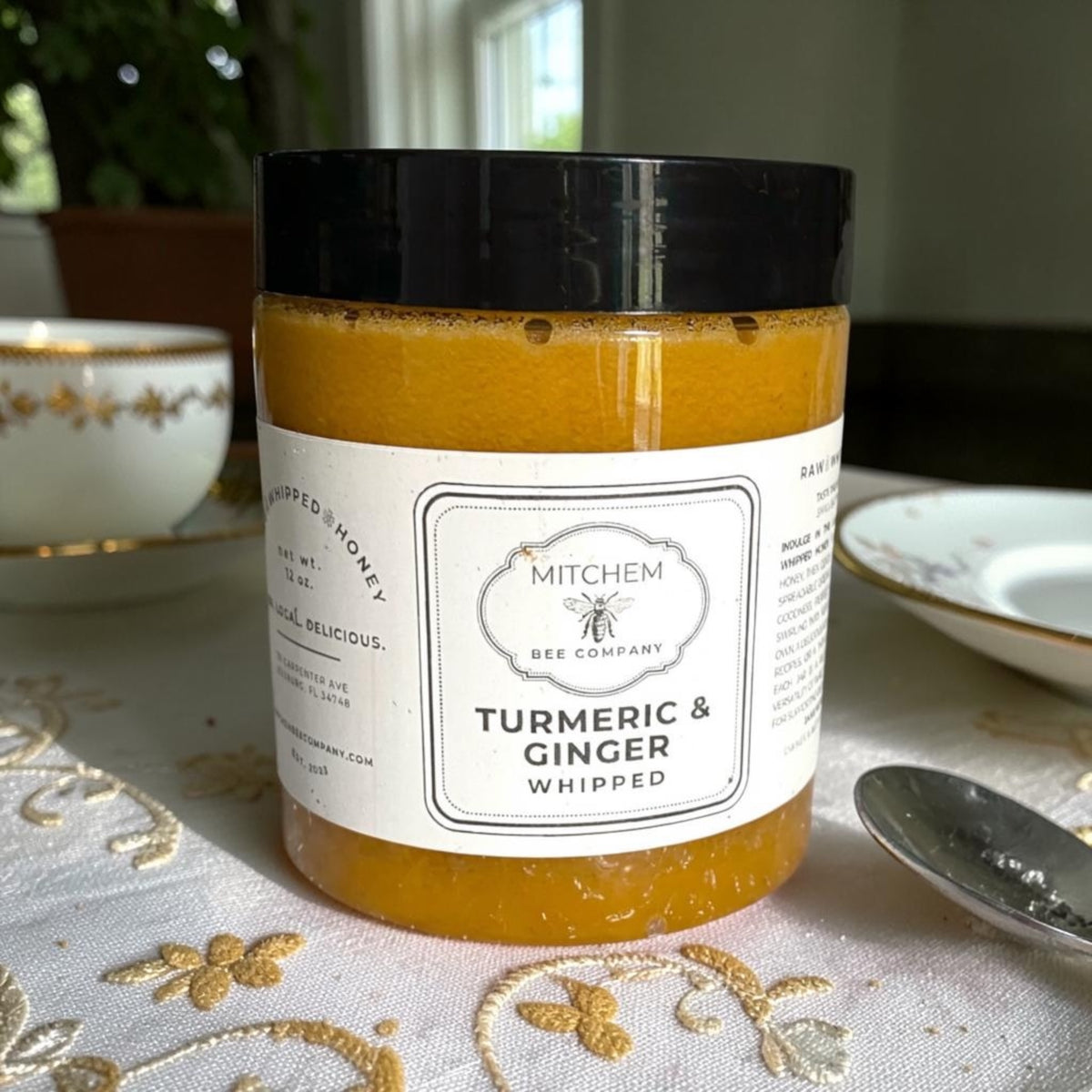 A jar of Mitchem Bee Company Turmeric & Ginger Whipped Honey sits on a white embroidered tablecloth with gold detailing, surrounded by a teacup, spoon, and plate in a softly lit indoor setting.