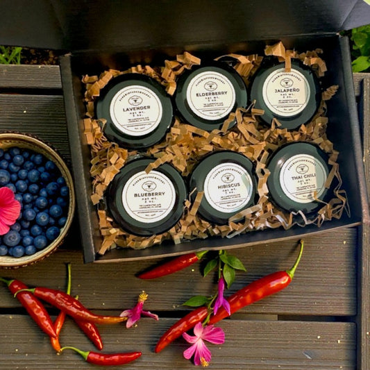 Set of small glass honey containers with labels in a decorative box, surrounded by chili peppers and flowers on a wooden surface.