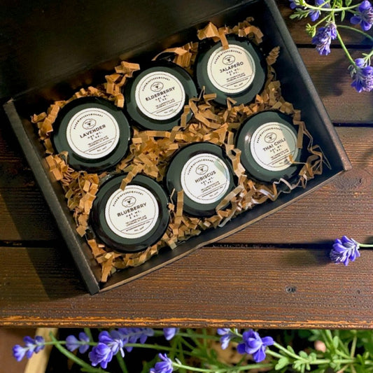Set of six small honey containers with labels in a decorative box on a wooden surface with lavender flowers.