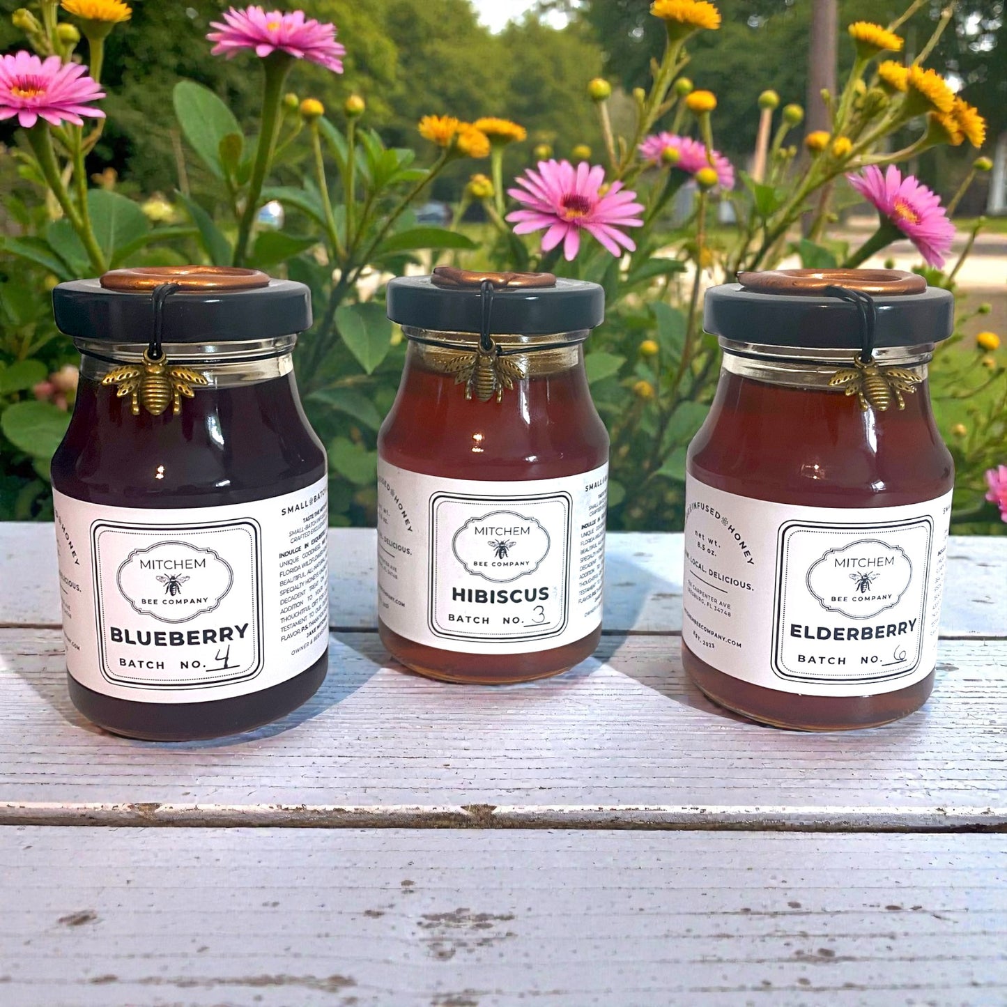 Three jars of infused honey labeled Blueberry, Hibiscus, and Elderberry on a wooden surface with flowers in the background.