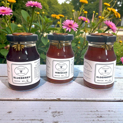 Three jars of infused honey labeled Blueberry, Hibiscus, and Elderberry on a wooden surface with flowers in the background.