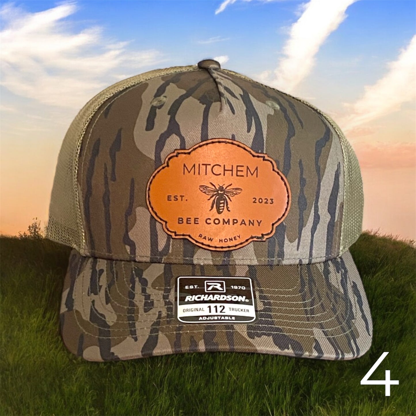 Brown Green Camouflage snapback hat with a Mitchem Bee Company branded leather patch on grass with a sunset background