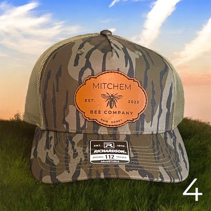 Brown Green Camouflage snapback hat with a Mitchem Bee Company branded leather patch on grass with a sunset background
