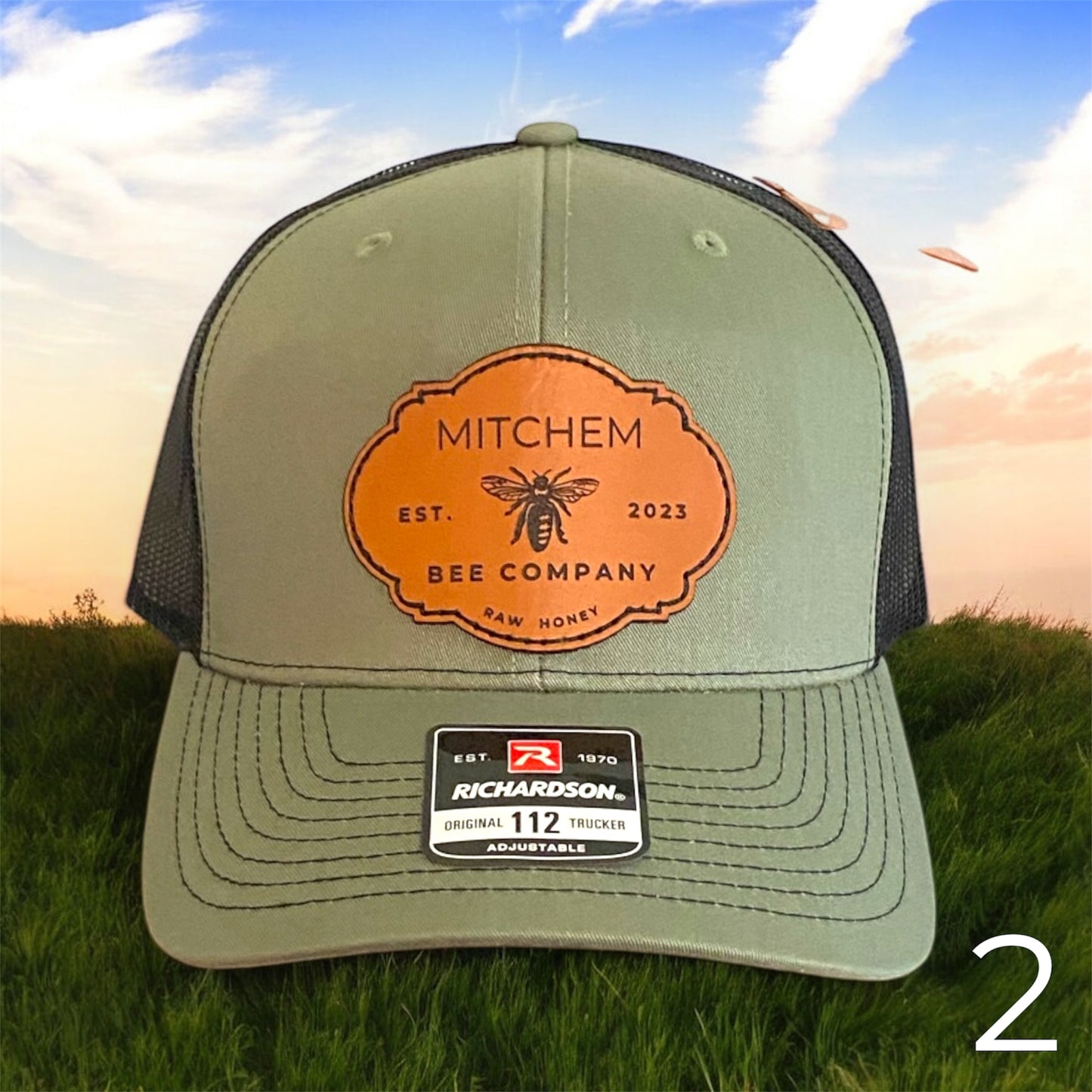Green cap with brown leather 'Mitchem Bee Company' logo patch with black mesh back on grass with sunset background
