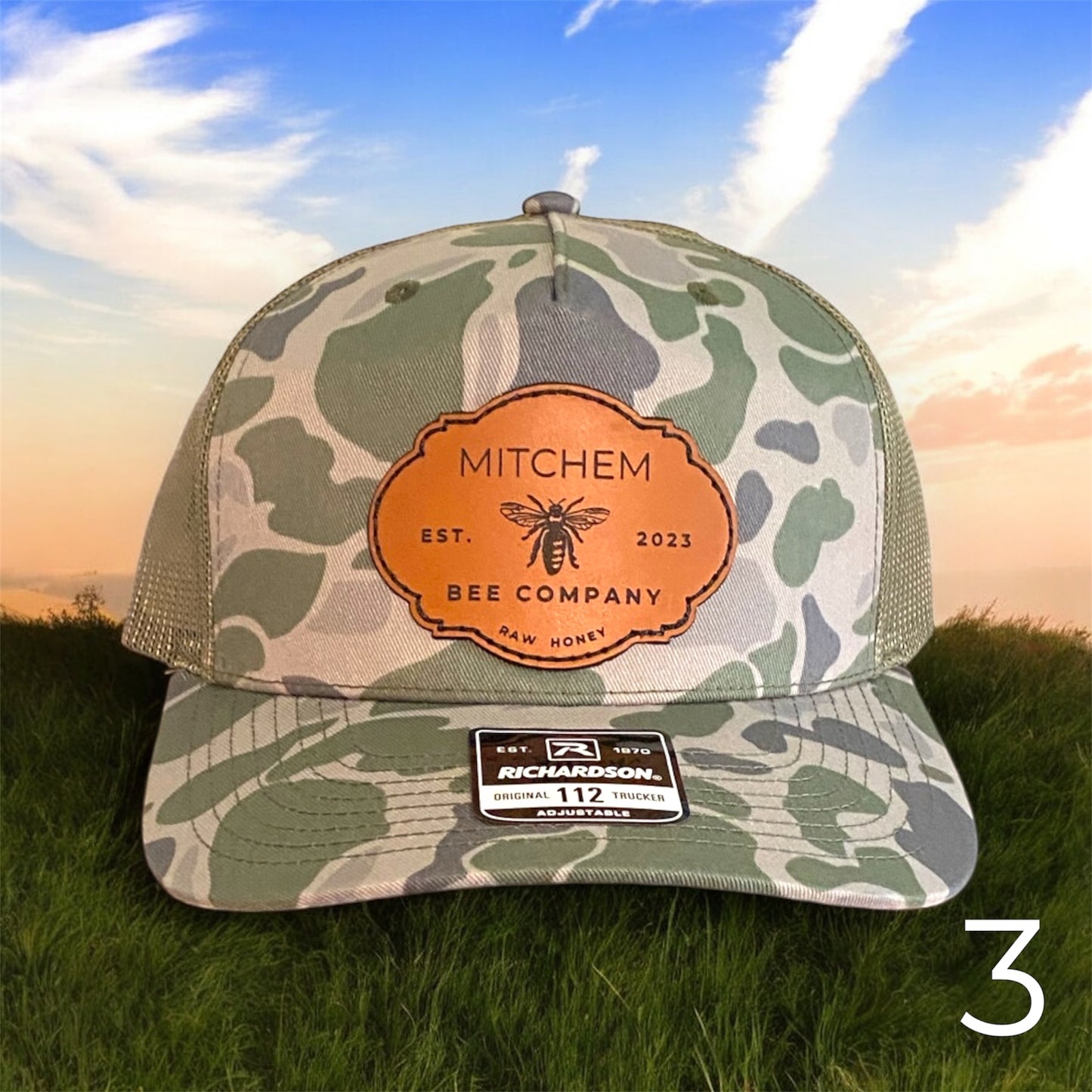Light green amouflage cap with a brown leather Mitchem Bee Company patch on grass with a sunset background