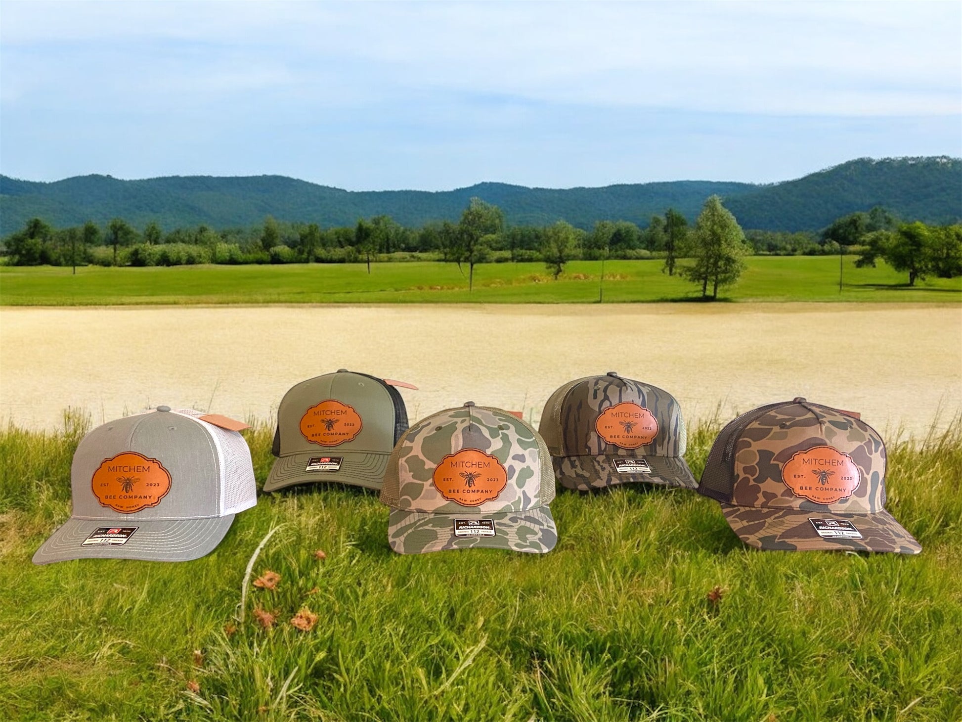 Five snapback baseball caps in gray, green, and 3 camouflage patterns featuring leather Mitchem Bee Company patches on a grassy field with mountains in the background.