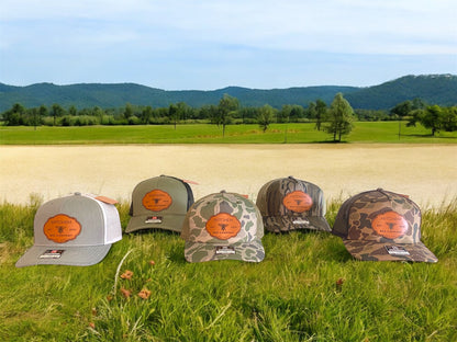 Five snapback baseball caps in gray, green, and 3 camouflage patterns featuring leather Mitchem Bee Company patches on a grassy field with mountains in the background.