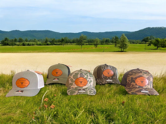 Five snapback baseball caps in gray, green, and 3 camouflage patterns featuring leather Mitchem Bee Company patches on a grassy field with mountains in the background.