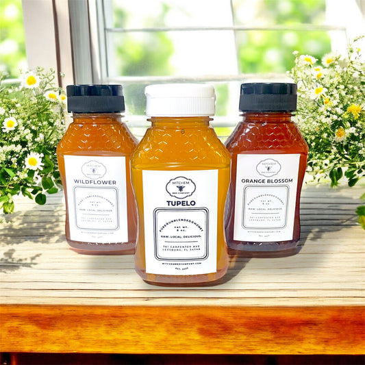 Three 8 oz. raw honey bottles with Mitchem Bee Company labels on a wooden surface with a window and flowers in the background