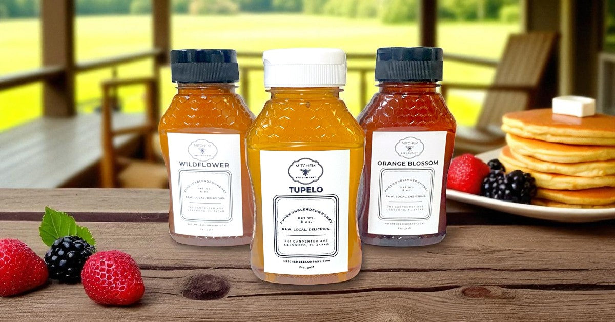 Three bottles of Mitchem Bee Company raw, local honey trio with labels on a wooden surface with berries and pancakes in the background