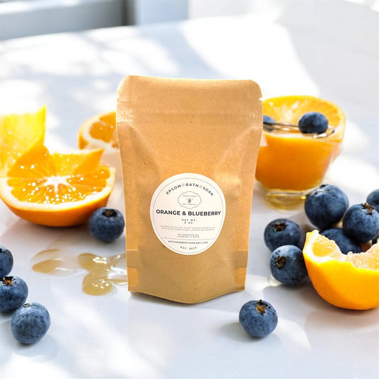 Packaged Epsom Bath Soak labeled 'Orange & Blueberry' with oranges, blueberries, and a glass of juice on a white surface.