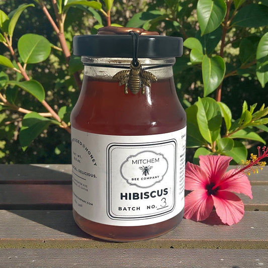 Jar of Mitchem Bee Company hibiscus honey with a bee decoration, surrounded by green leaves and a pink flower.
