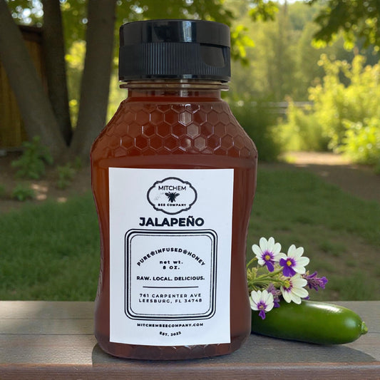 Bottle of Mitchem Bee Company Jalapeño honey with a label, surrounded by flowers and a jalapeño on a wooden surface with a natural background.