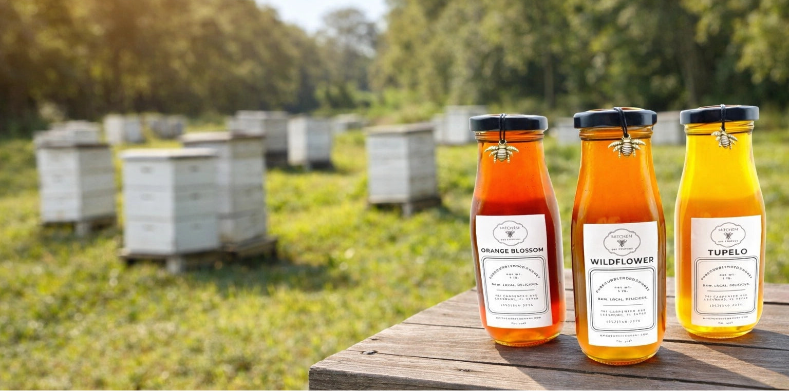 Three glass bottles of Mitchem Bee Company honey on a wooden table with bee hives in the background.